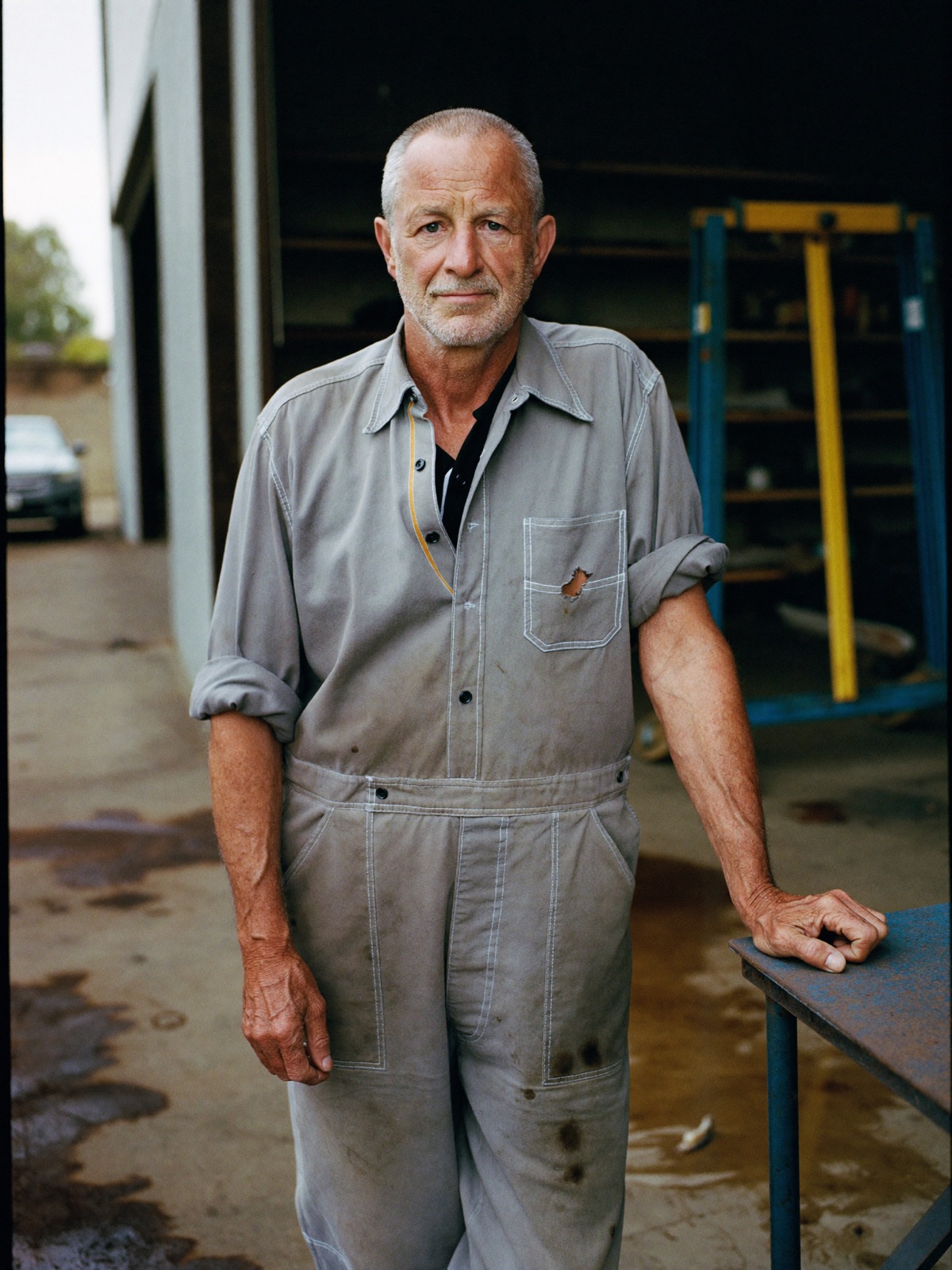 Portrait of a mechanic in worn stone-grey overalls standing at the open door of a workshop.
